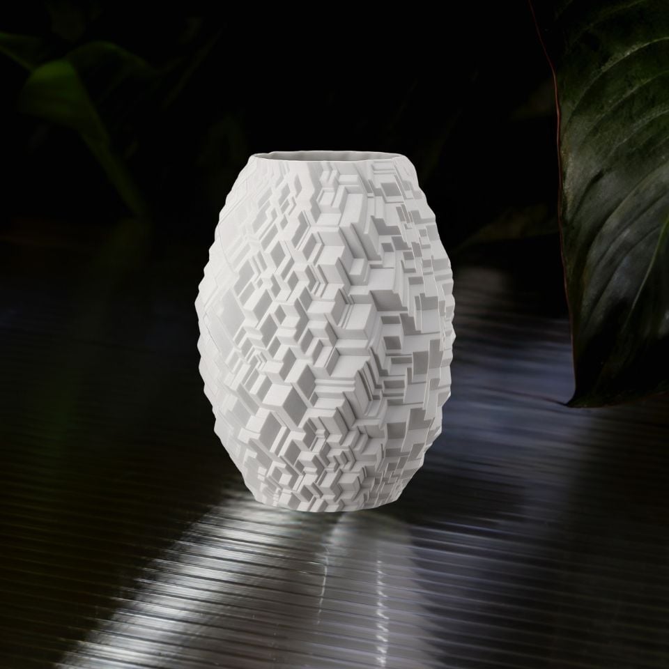 A white porcelain vase with intricate 3D geometric patterns, placed on a glossy surface with dark green foliage.