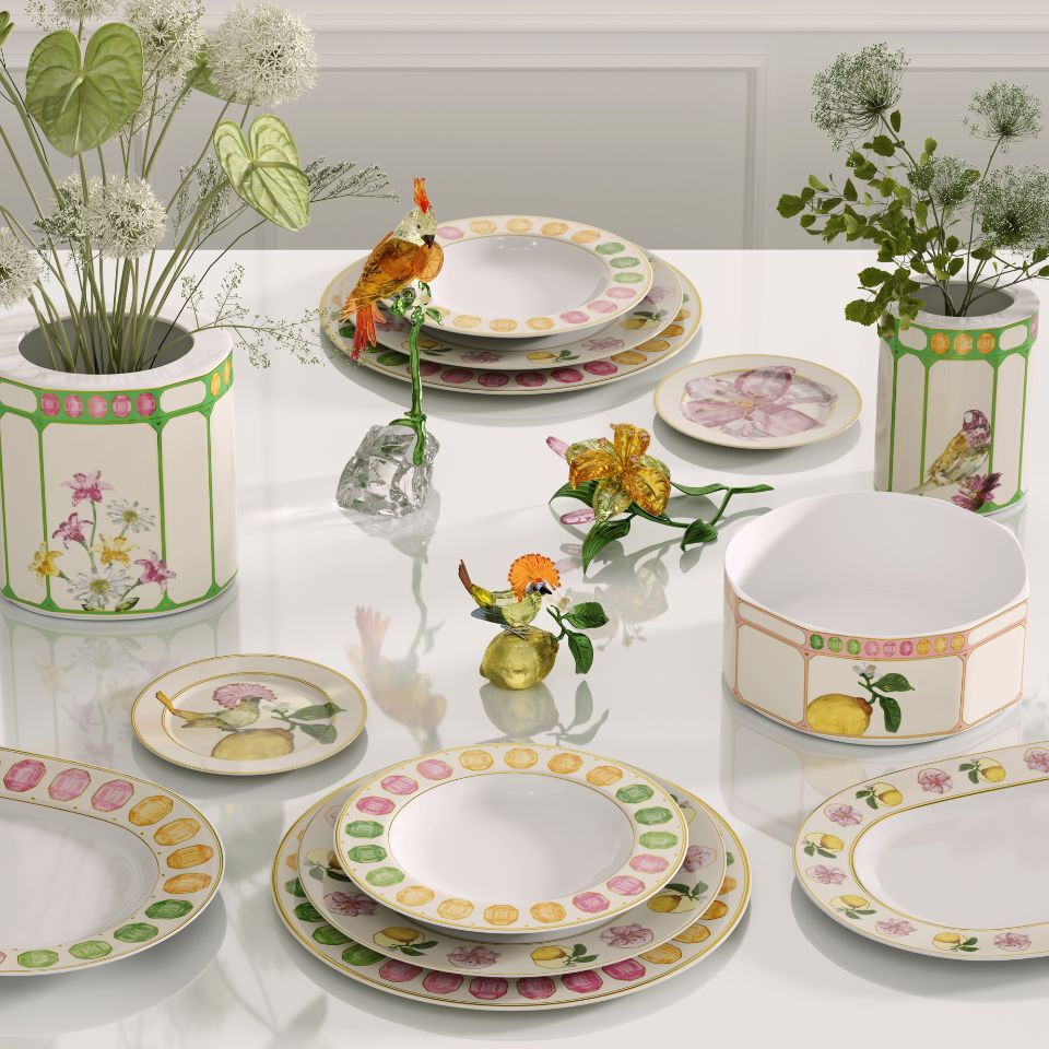 Whimsical Idyllia porcelain collection by Swarovski x Rosenthal with delicate botanical and fruit illustrations in soft pastel tones, accented with crystal bird and flower sculptures, arranged on a glossy white table for a poetic tablescape.