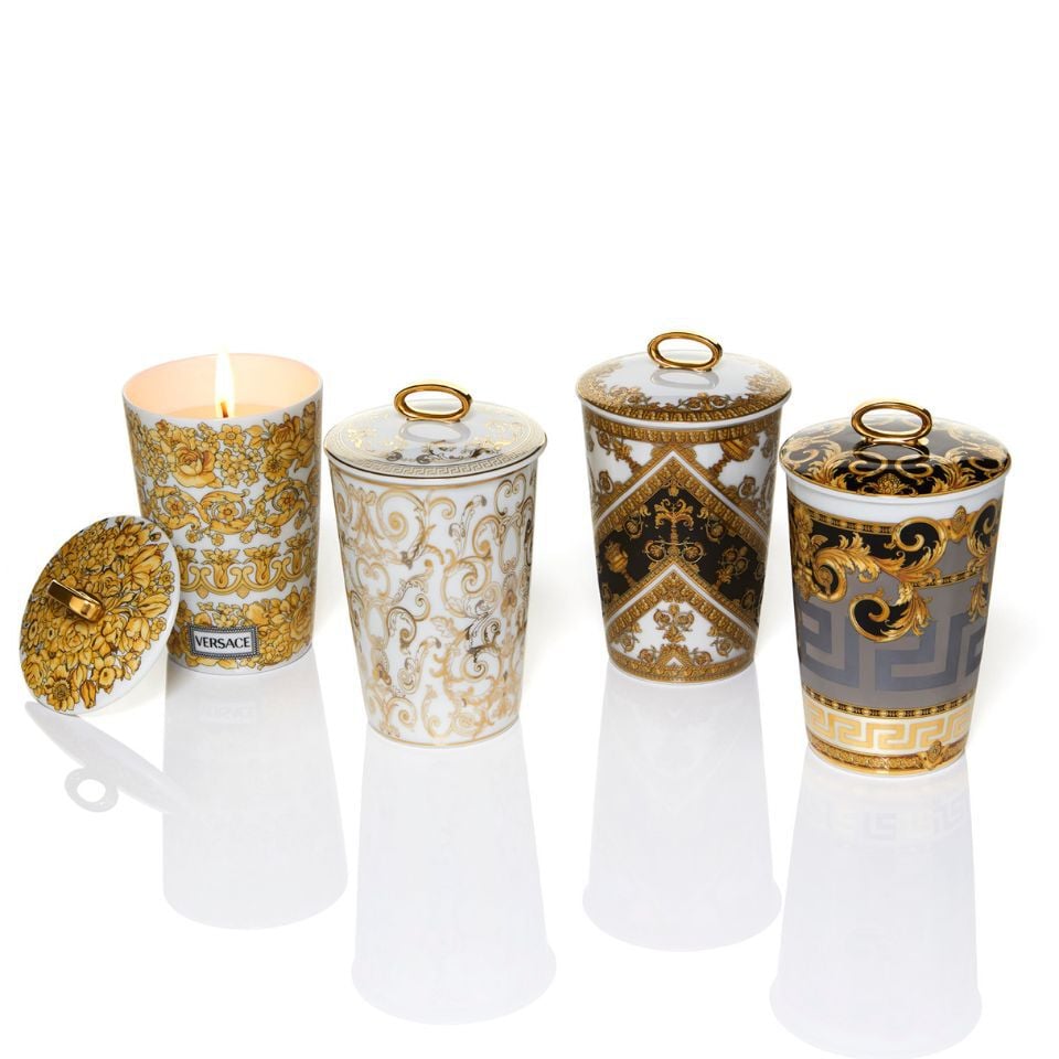 Elegant Versace scented candles in porcelain containers with lids, decorated in classic gold baroque patterns, including one open candle with a warm flame.