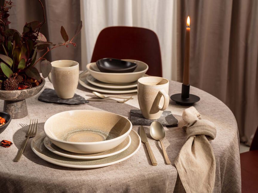Tavola apparecchiata con stoviglie in gres di Junto Rosenthal: piatti, ciotole e tazze nei toni naturali, abbinati a candele, posate e tovaglioli di stoffa.