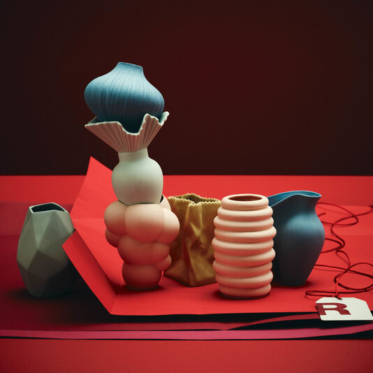 Farbige Minivasen nebeneinander aufgereiht und teilweise gestapelt stehen auf dunkelrotem Geschenkpapier