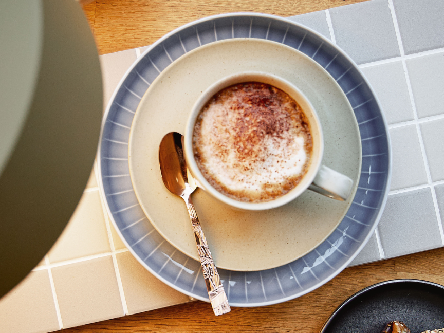 Tasse &agrave; caf&eacute; Rosenthal de forme classique l&eacute;g&egrave;rement conique, avec une anse filigran&eacute;e et une soucoupe assortie au bord structur&eacute;, remplie de cappuccino et dispos&eacute;e sur un plateau carrel&eacute; clair.