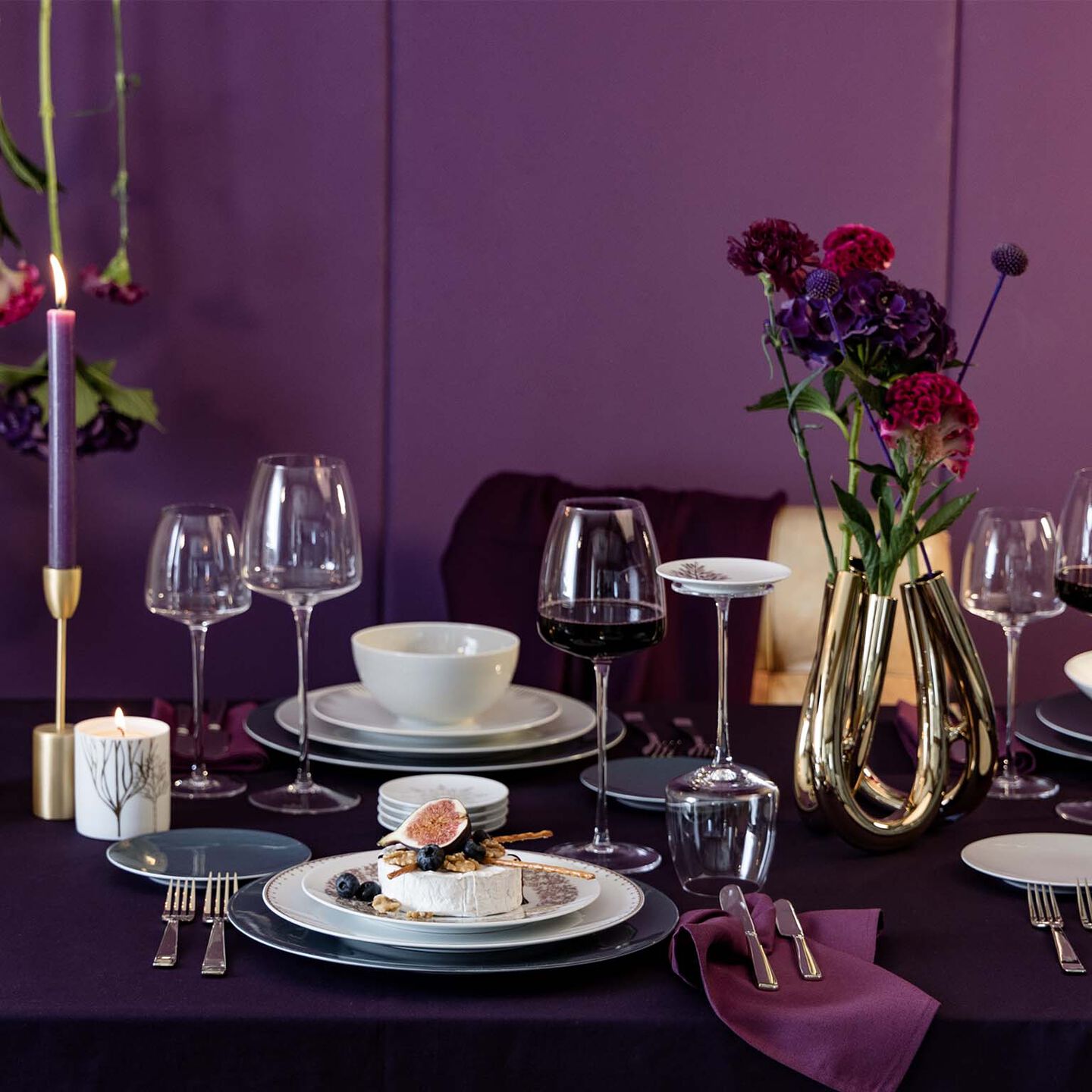 Elegantly set table with Rosenthal tableware and glassware against a purple backdrop, golden vase with flowers, and atmospheric candlelight.