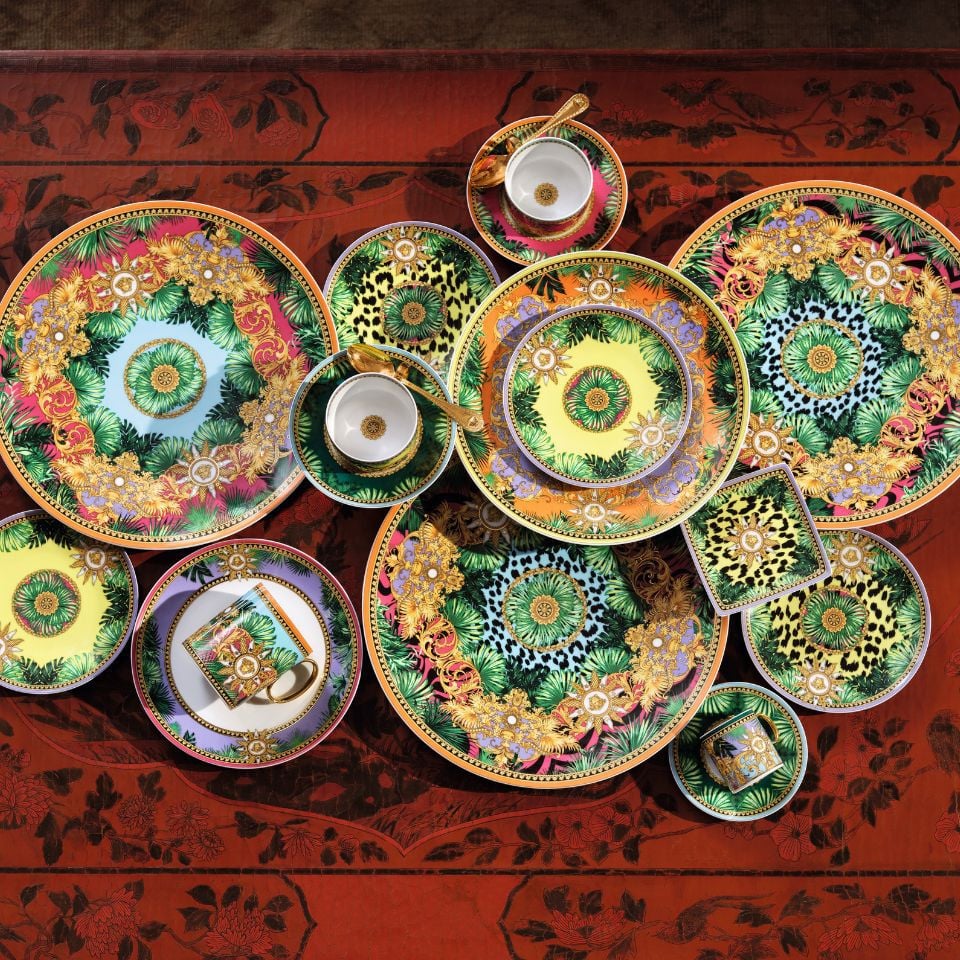 Bold and colorful Jungle Animalier dinnerware set by Versace, with tropical motifs, animal prints, golden flowers, and lush greenery on a red patterned table.