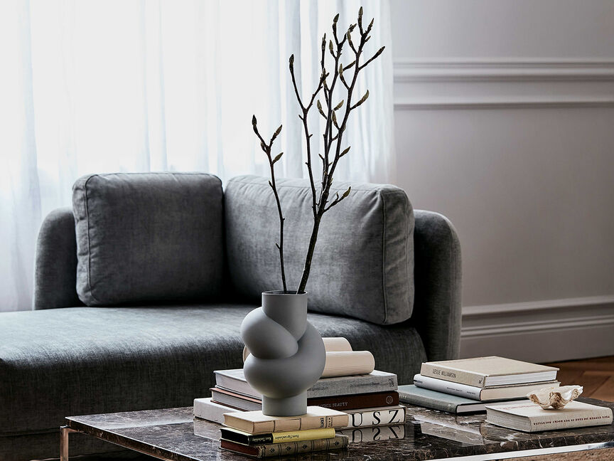 Node Lava für Ihr Zuhause Vase Node Lava mit Zweig steht auf einem Bücherstapel auf einem Marmortisch vor einem grauen Sofa.