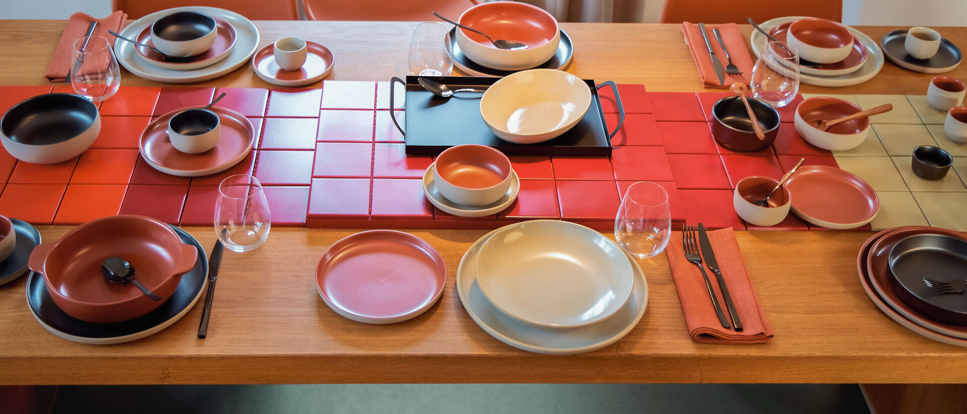 A wooden table laid with colourful tableware in terracotta, red and ros&eacute; &ndash; bowls with lids, plates and glasses on red placemats, surrounded by orange chairs.