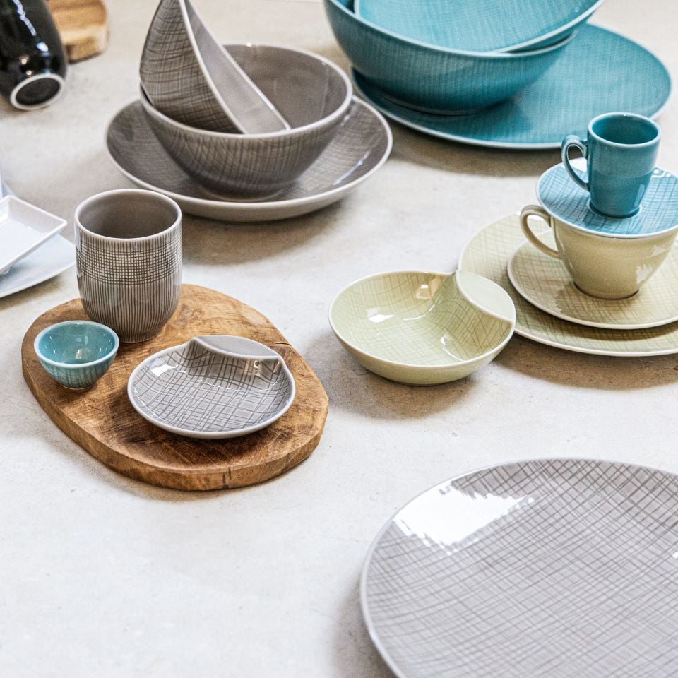 A casual mix of ceramic bowls, cups, and plates in soft grey, green, and turquoise tones with fine textured patterns.