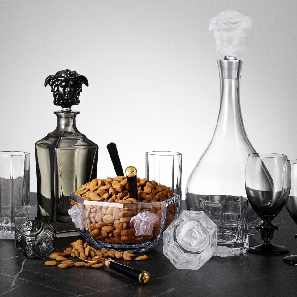 Versace Medusa Lumière crystal barware set including decanters, tumblers, and a bowl filled with almonds, each adorned with sculpted Medusa reliefs and paired with black and gold accents.