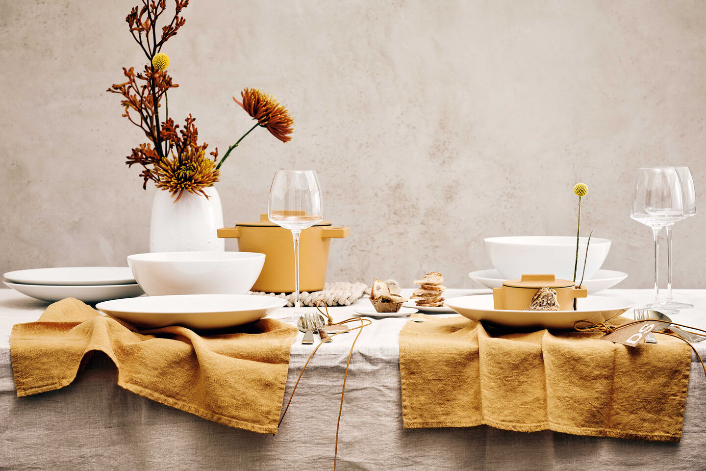Rosenthal TAC Dining Collection mit dem neuer neuen TAC Allround Schüssel auf einem gedeckten Tisch mit gelben Stoffservietten, Weingläsern und Herbstblumen in einer Vesi Vase.