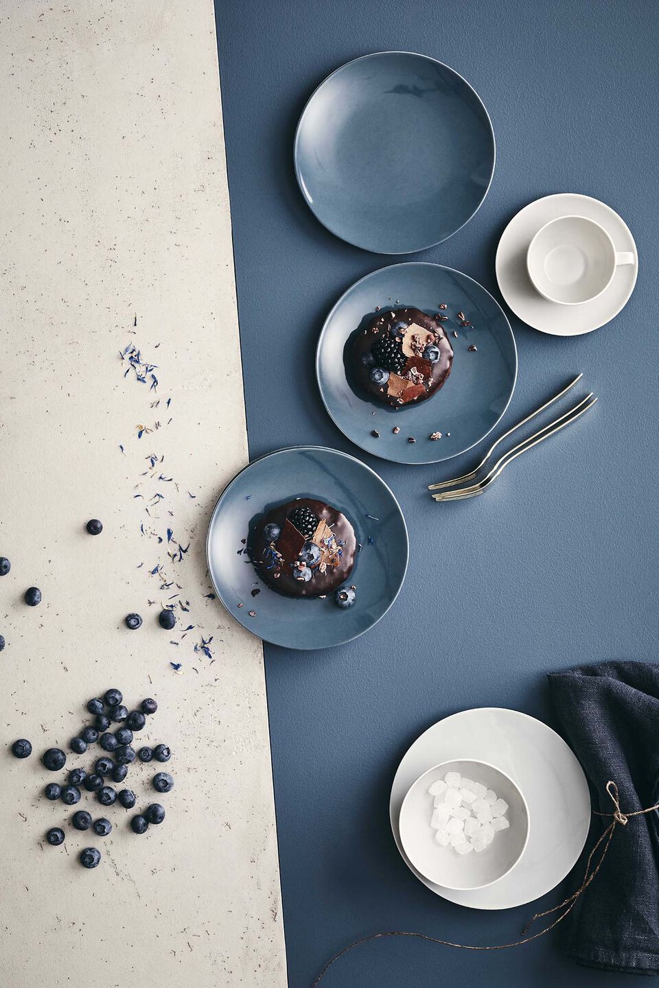 Elegant TAC Sensual View from above of a concrete-coloured and blue background on which plates and bowls from TAC Sensual in Comfort Blue and Gentle Grey are arranged in a jumble, some with desserts of chocolate and berries.
