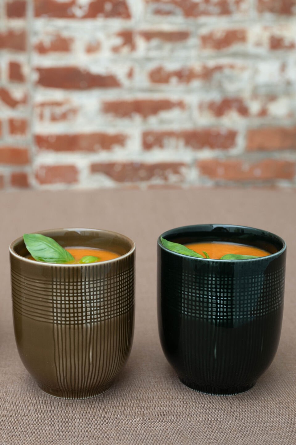 Two MUCA cups, filled with soup and garnished with a basil leaf, stand in front of a brick-look background.
