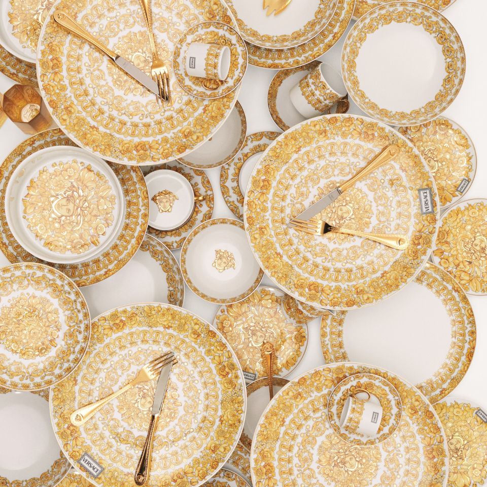 Opulent Medusa Rhapsody porcelain dinnerware by Versace with lush golden floral baroque designs, layered in an artistic arrangement with golden cutlery on white background.