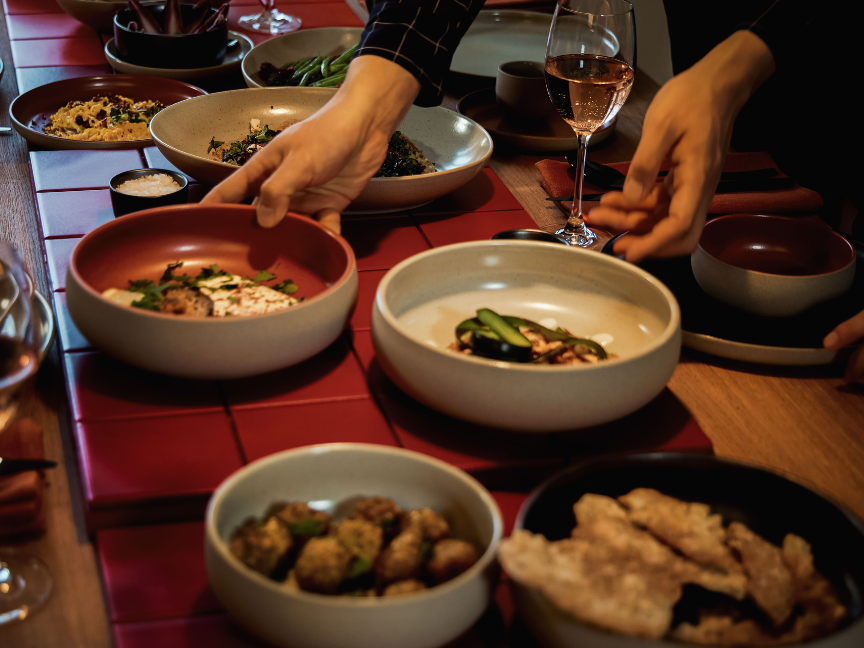 People are passing around Rosenthal bowls filled with vegetable dishes across a laid table with red placemats, wine glasses and other accompaniments.