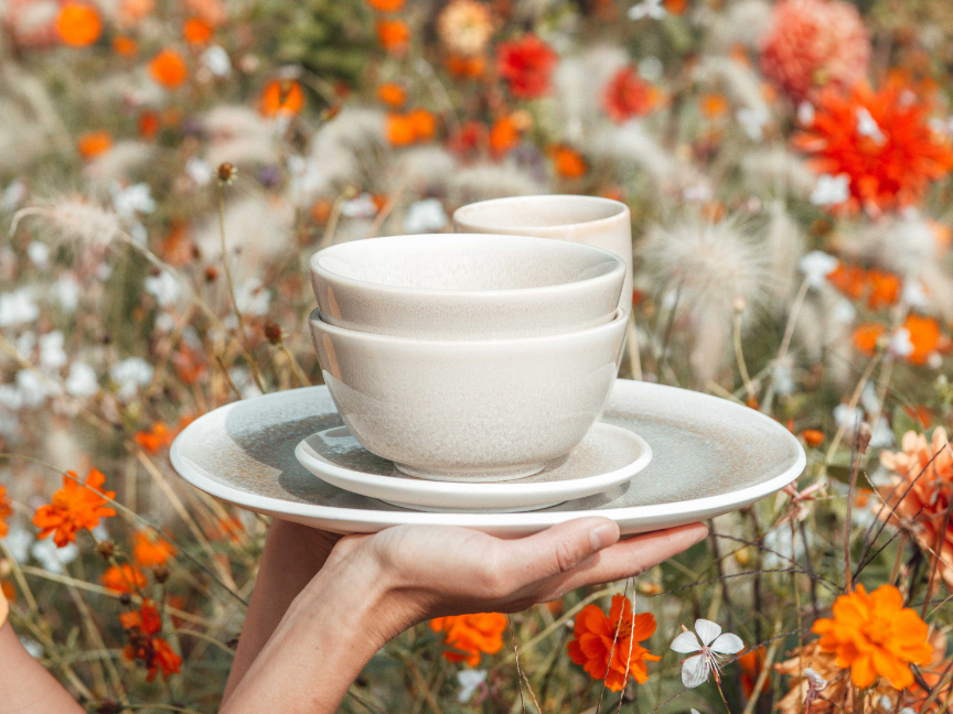 Rosenthal tableware in cream-coloured ceramic, comprising plates, bowls and cups, attractively displayed against a backdrop of a summer meadow in bloom with orange flowers.