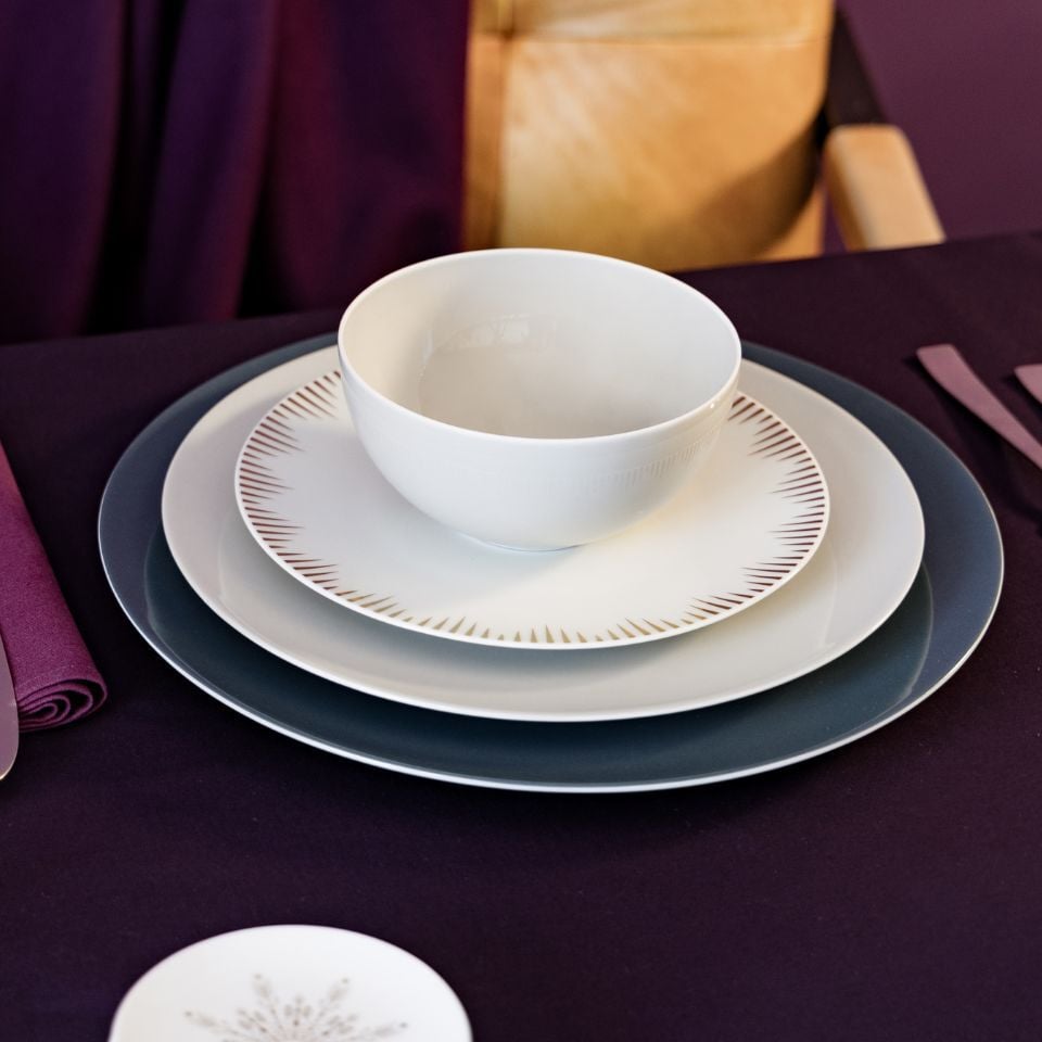 A layered setting with matte plates in white and dark grey, and a central plate with bronze sunburst detailing.