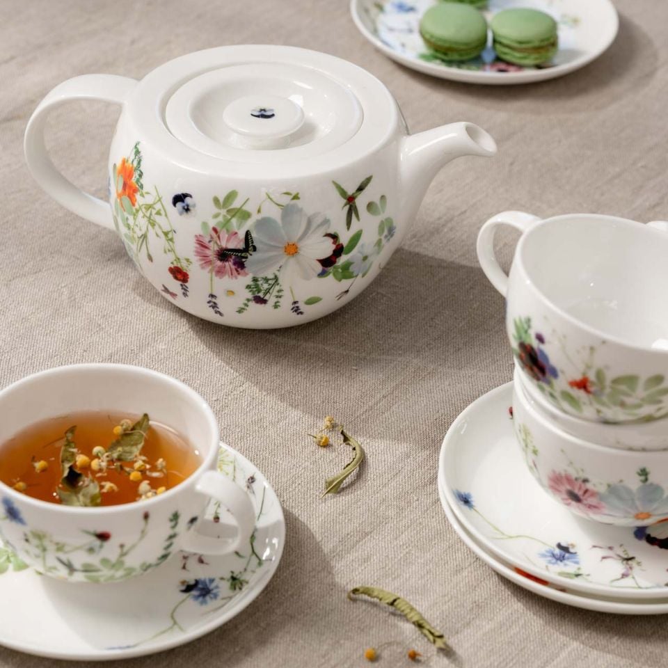A white porcelain teapot and teacups decorated with colorful flower and leaf motifs, arranged on a beige linen tablecloth with a cup of herbal tea and a plate of green macarons.