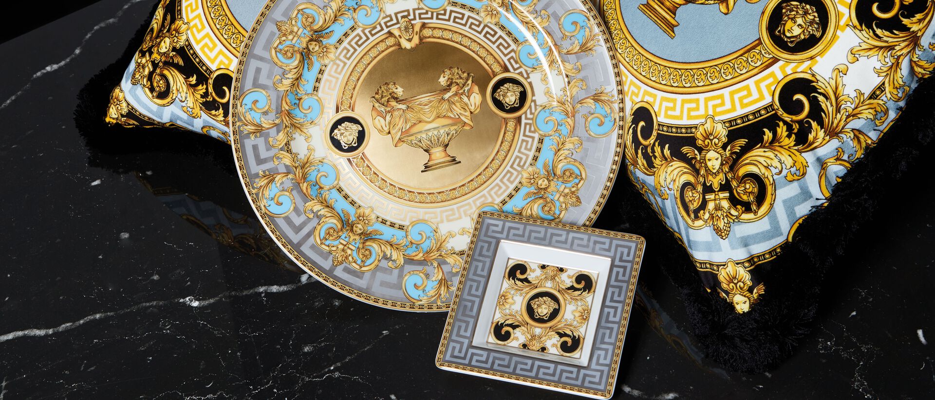 Versace Medusa von Rosenthal Teller und Schalen in Hellblau, Gold und Wei&szlig; mit opulentem Barockdekor, Medusa-Relief und zentralem L&ouml;wenmotiv, arrangiert auf schwarzer Marmoroberfl&auml;che mit passender quadratischer Schale.