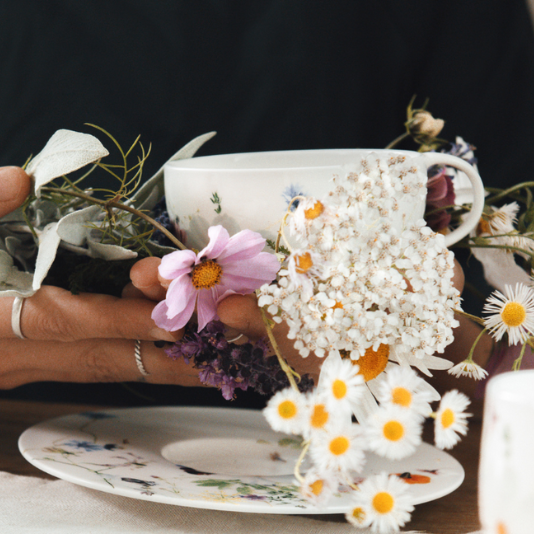 Hände halten eine Rosenthal-Tasse mit zartem Blumenmuster, umgeben von Wiesenblumen wie Kamille und Schleierkraut.