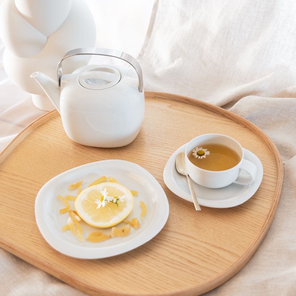 A bright tray with a white teapot, chamomile tea, and a plate of lemon slices and candied zest on soft fabric.