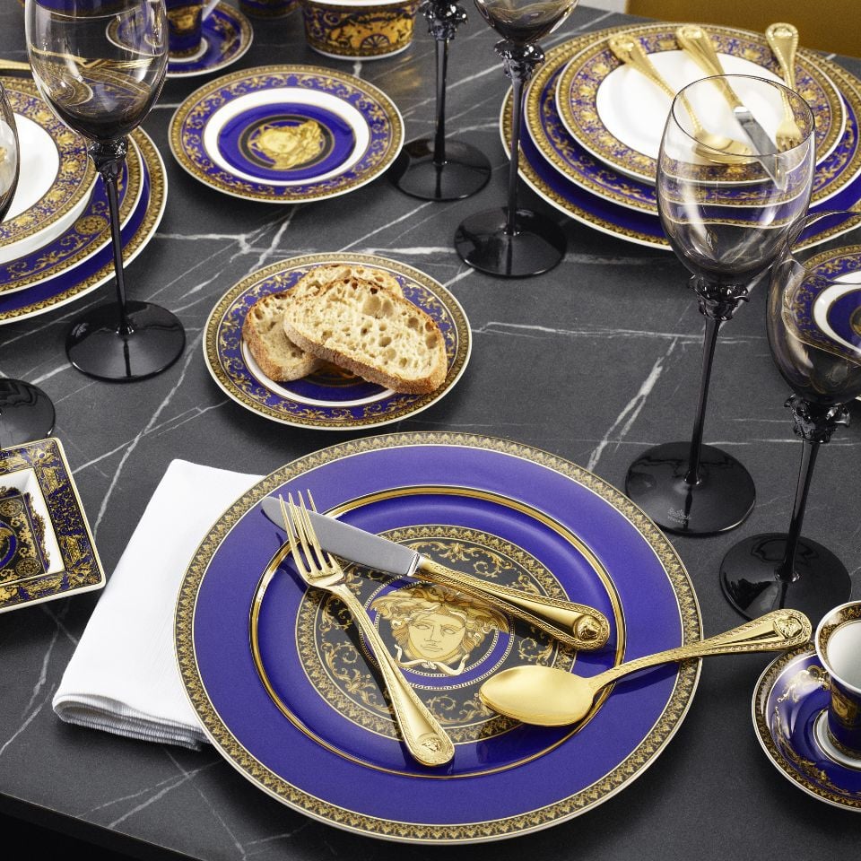 Elegant Versace tableware from the Medusa Blue collection, featuring rich cobalt blue and gold plates adorned with the iconic Medusa head, arranged on a dark marble table.
