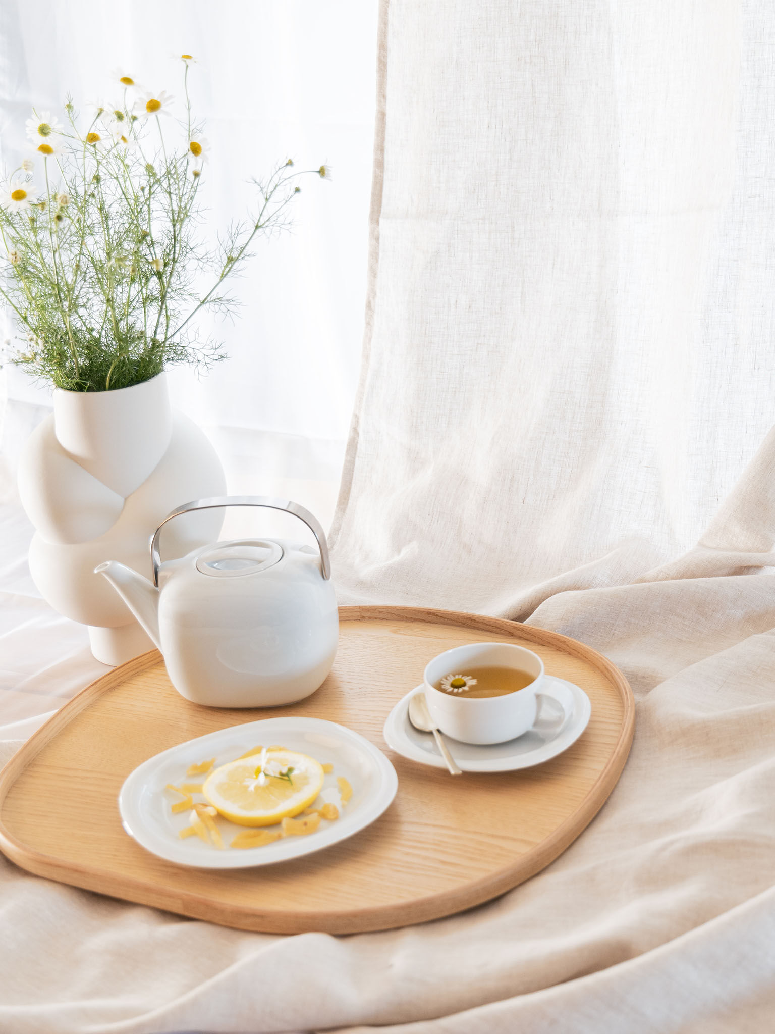 Suomi teapot, plate and filled teacup with saucer on a round wooden tray with Node vase and linen cloth in the background.