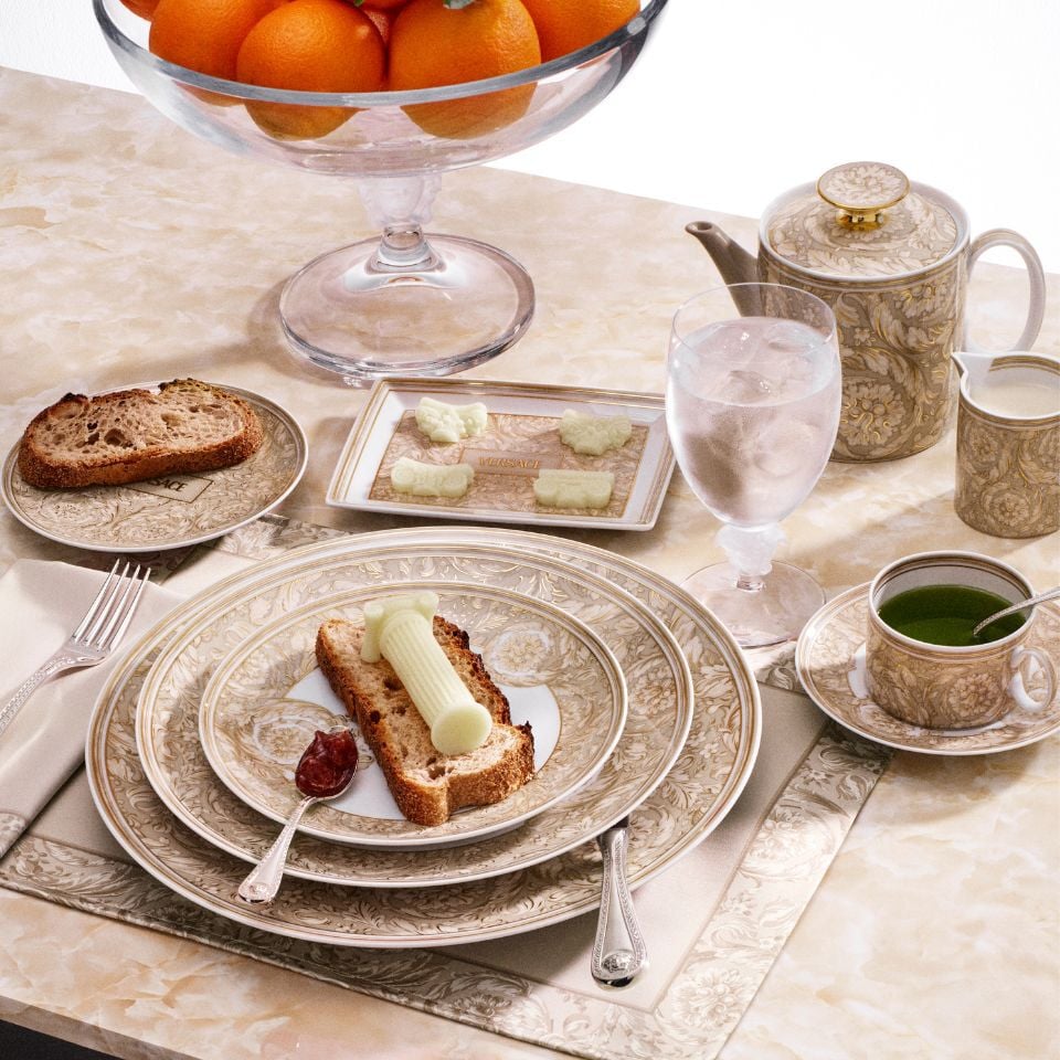 Luxurious Barocco porcelain breakfast set by Versace in soft gold tones, with ornate patterns, butter on toast, green tea, and a glass of water on a marble table.