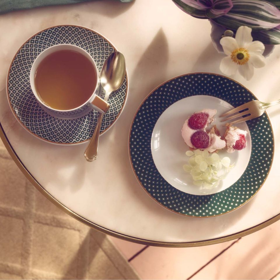 A small round table with a cup of tea, a dark green plate with a pastry topped with raspberries, a golden fork and spoon, and a white flower placed nearby.