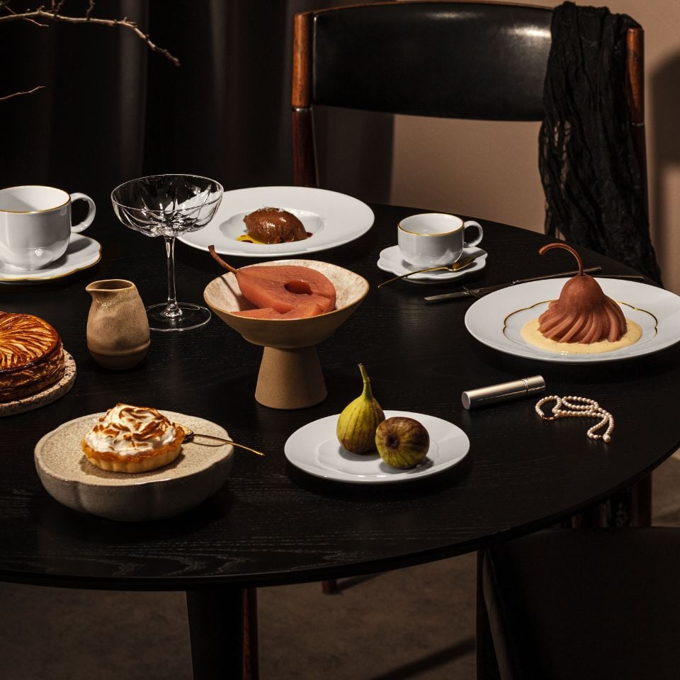 A dark table with artistic desserts, figs, and minimalist white plates, styled with pearls and moody lighting.