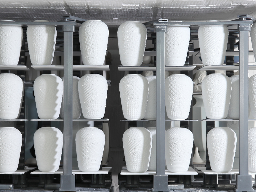 Porzellanherstellung: Wei&szlig;e, ungebrannte Vasen stehen in Reihen auf einem industriellen F&ouml;rdergestell in einer Produktionsanlage.