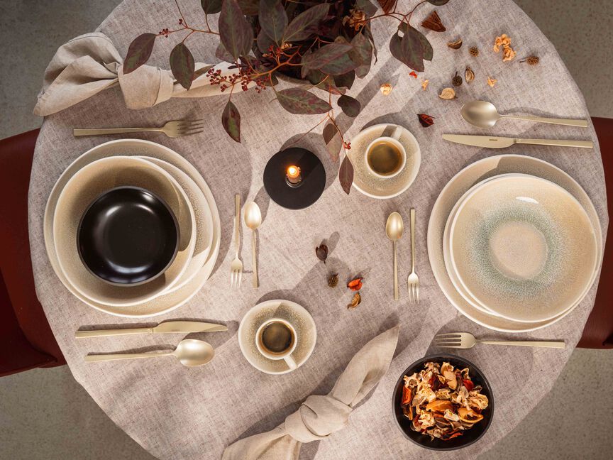 Tavolo rotondo con stoviglie in gres di Rosenthal: piatti, ciotole e tazze in tonalit&agrave; naturali, completati da candele, posate e tovaglioli in tessuto.