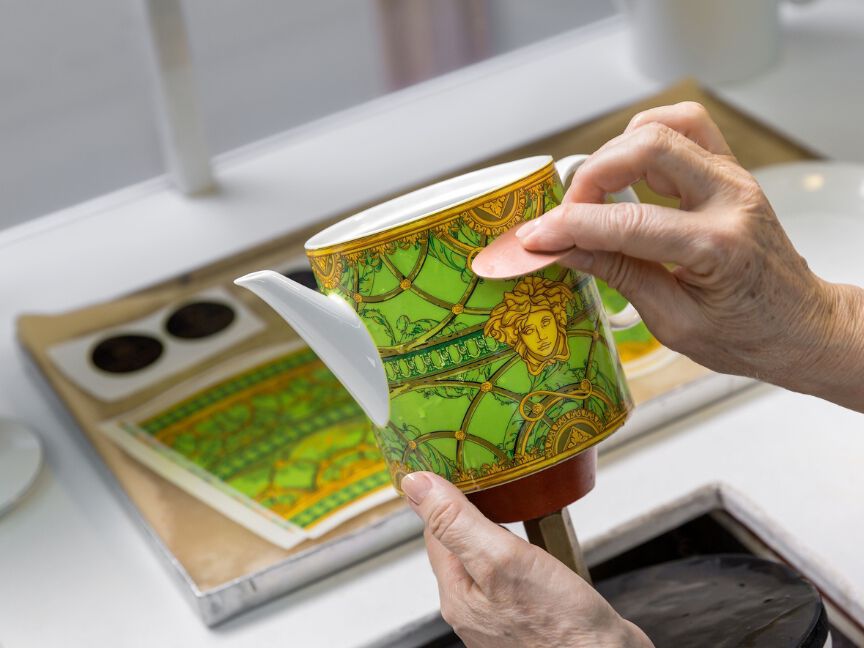 Porzellandekoration: Eine Person tr&auml;gt vorsichtig ein farbiges Design von Hand auf eine wei&szlig;e Teekanne auf.