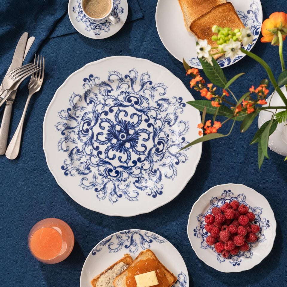 A breakfast table with blue and white porcelain plates featuring floral patterns, raspberry bowl, toast, and espresso on a dark blue tablecloth.