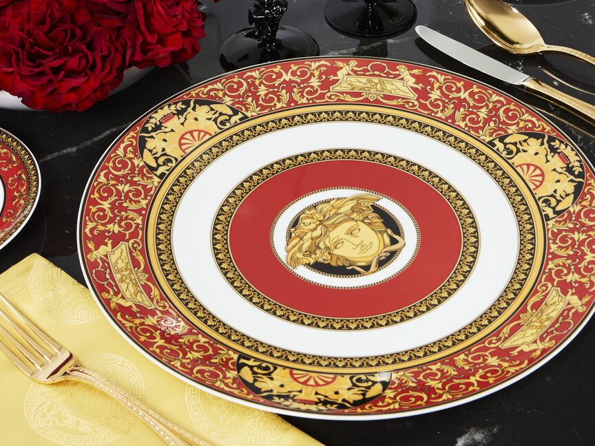 Versace Medusa von Rosenthal Speiseteller in Rot, Gold und Wei&szlig; mit opulentem Barockdekor und zentralem Medusa-Motiv, inszeniert auf schwarzer Tischoberfl&auml;che mit goldfarbenem Besteck, gelber Serviette und roten Rosen.