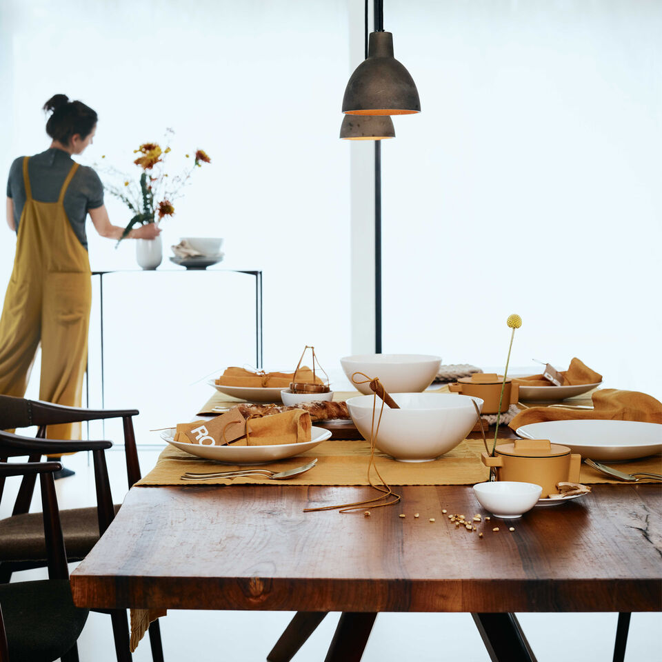 Side view of a table set with TAC White with yellow napkins, tablecloths, the TAC Allround bowl in the centre of the table and a woman in yellow dungarees in the background draping yellow flowers in a vase.
