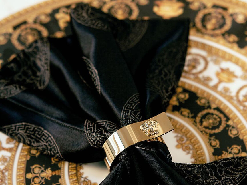 Versace Medusa von Rosenthal Speiseteller in Schwarz und Gold mit opulenter Barockbord&uuml;re, inszeniert mit schwarzer Stoffserviette und goldfarbenem Serviettenring mit Medusa-Emblem auf gedecktem Tisch.