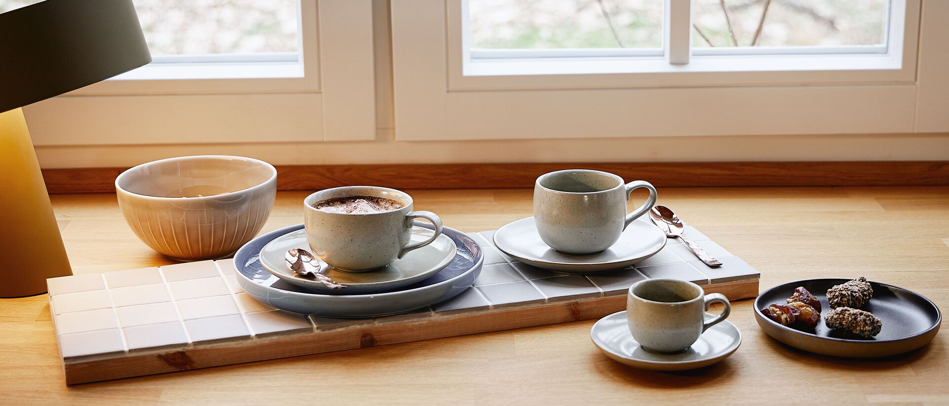 Tasses &agrave; caf&eacute; de diff&eacute;rentes formes de Rosenthal, dont des tasses bomb&eacute;es et cylindriques avec soucoupes, dispos&eacute;es sur un plateau en bois avec une surface carrel&eacute;e dans un environnement de cuisine lumineux.