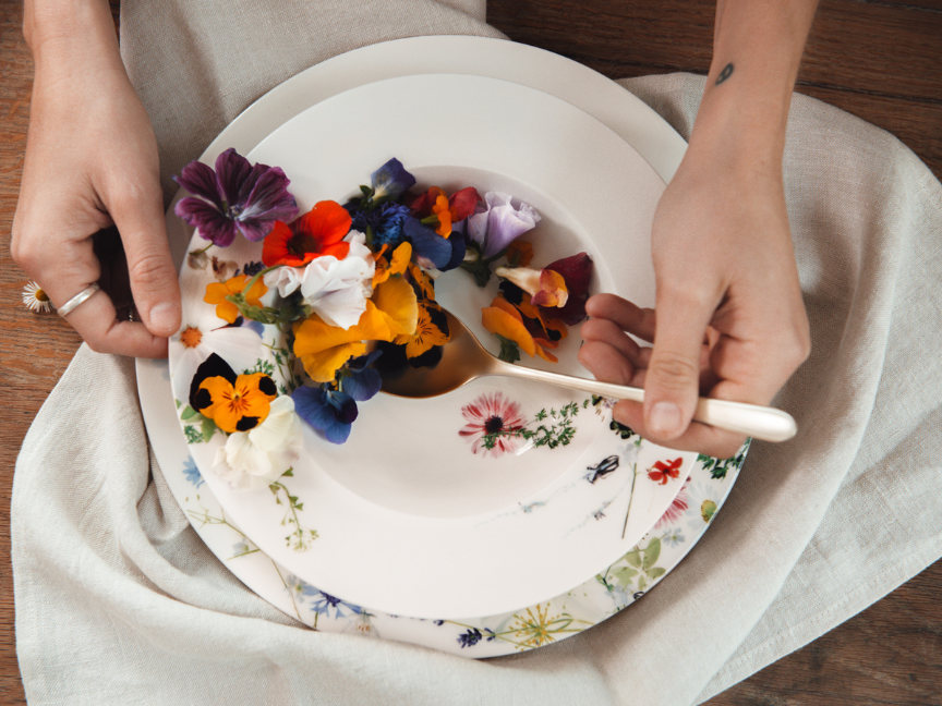 Hände halten einen Löffel über einem mit bunten essbaren Blüten dekorierten Porzellanteller mit floralen Motiven von Rosenthal Brillance.