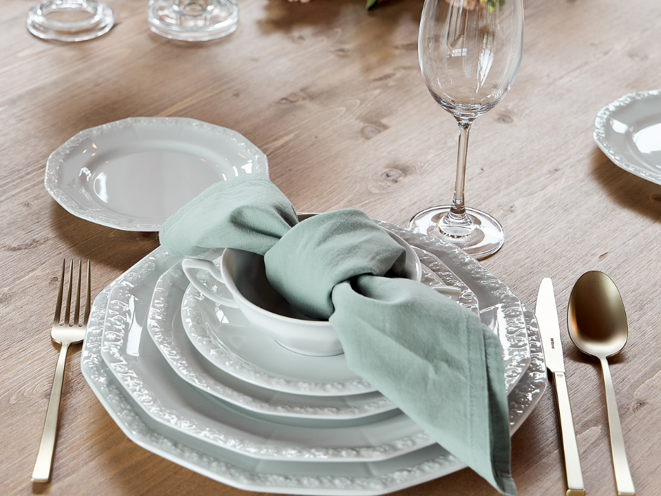 Festlich gedeckter Tisch mit weißem Rosenthal Maria Porzellan, hellgrüner Serviette, Goldbesteck und weißen Stumpenkerzen auf einem runden Holztisch