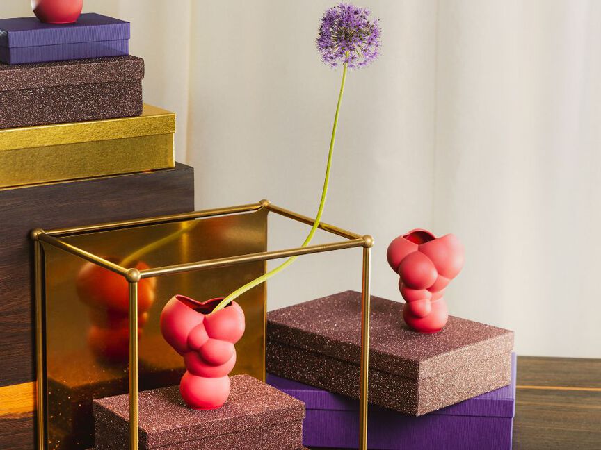 Dekorative Szene mit roten Designer-Vasen von Rosenthal auf glitzernden Geschenkboxen und einer lila Blüte in modernem Arrangement