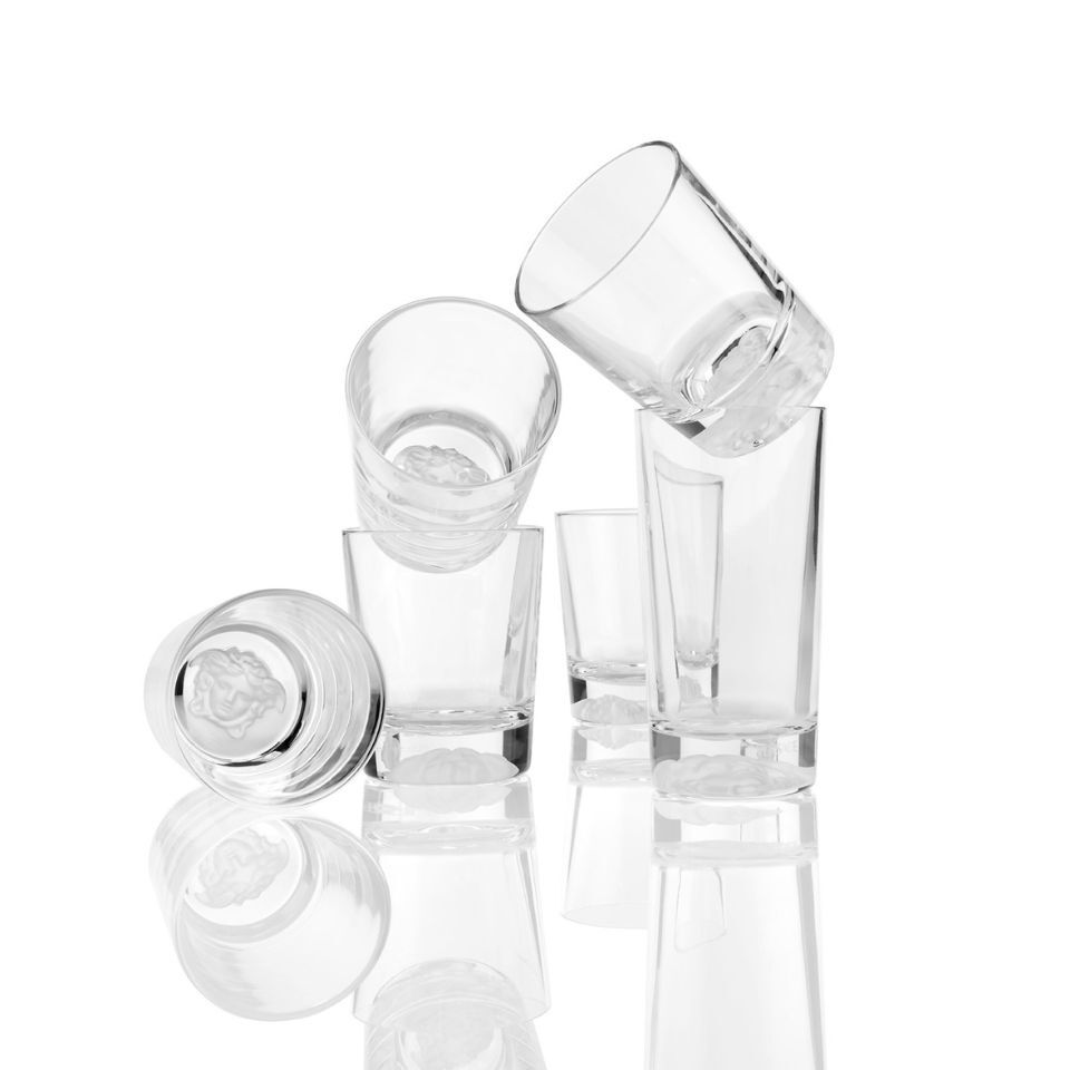 Set of stacked Versace crystal tumblers with embossed Medusa heads at the bottom, arranged playfully on a reflective white surface.