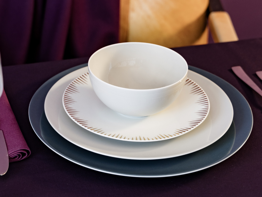 A modern table setting featuring Rosenthal&rsquo;s antipasti tableware. White bowls and plates with delicate decoration are arranged on dark blue placemats, creating an elegant contrast with the purple tablecloth.