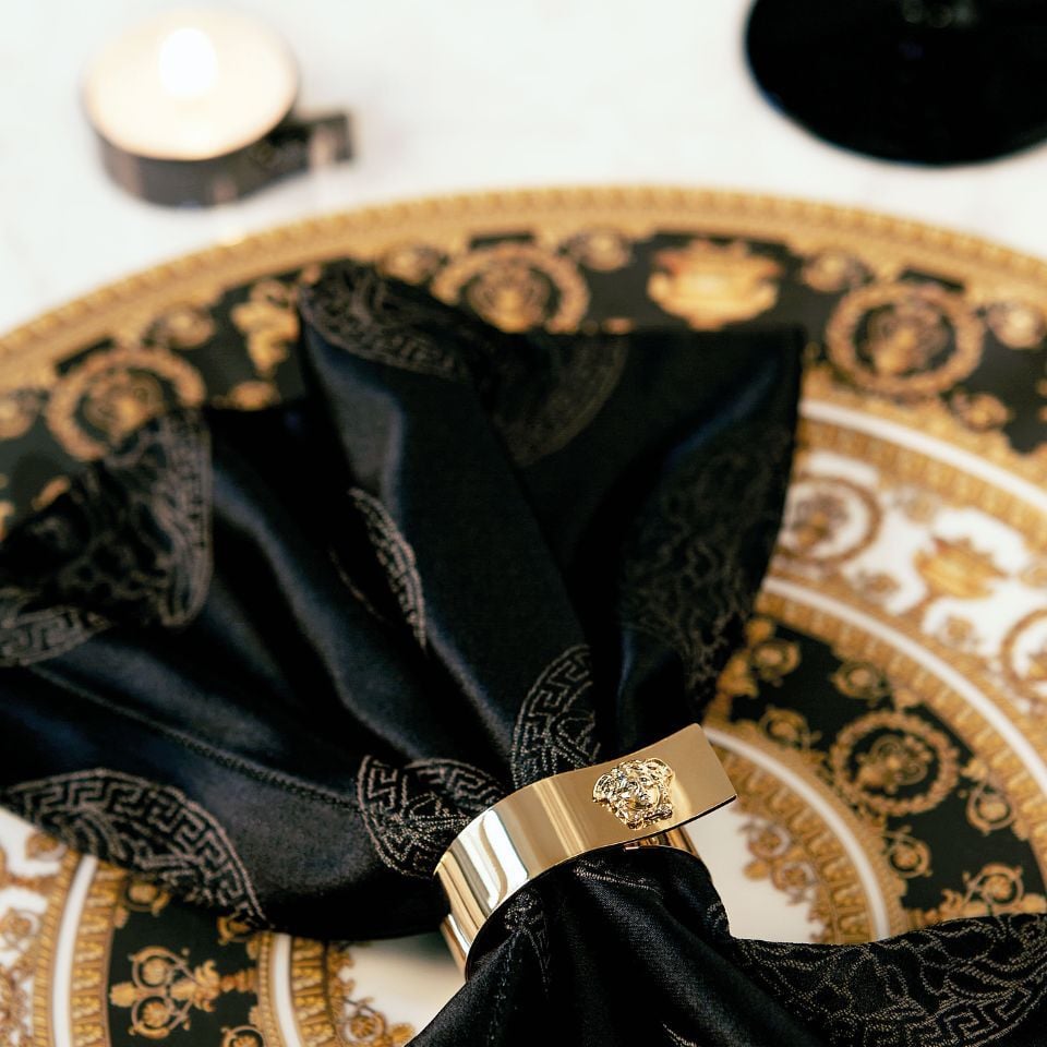 Close-up of a Versace table setting with black satin napkin held by a gold napkin ring featuring the Medusa emblem, placed on a black and gold baroque plate beside a candle.