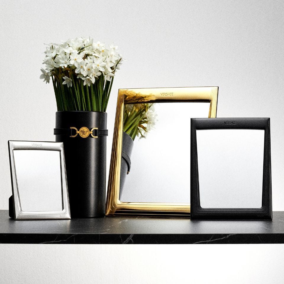 Versace luxury photo frames in silver, gold, and black finishes displayed on a dark shelf, accompanied by a black vase with white flowers, all embossed with the Versace logo.