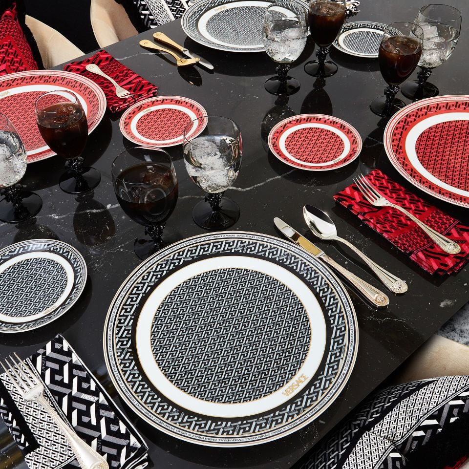 Versace’s La Greca Signature tableware in black and red geometric patterns, paired with crystal-clear black-stem glasses and coordinated napkins for a modern table setting.