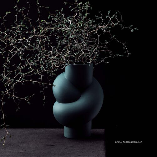 Node vase in the shade Abyss with branches against a black background and standing on a dark grey concrete surface.