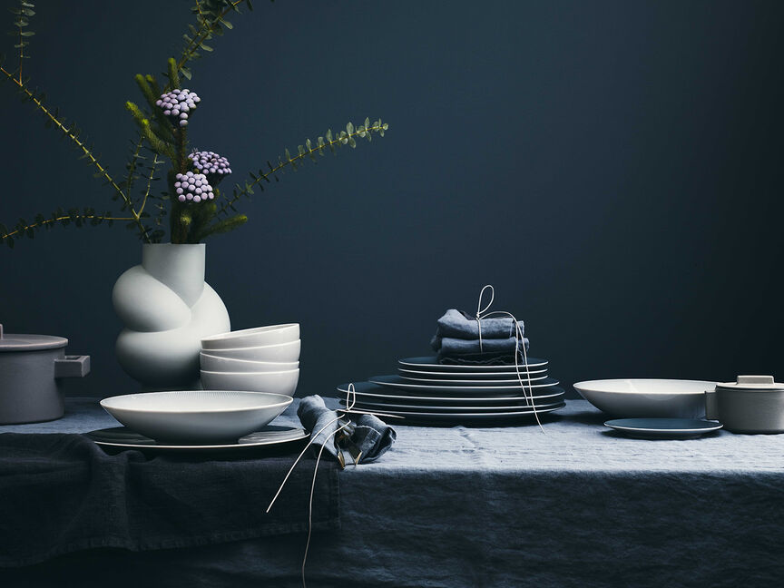 Perfect combination partners Linen tablecloth against a dark blue background with plates and bowls by TAC Sensual stacked on top and a white Node vase placed with flowers, sage and twigs.