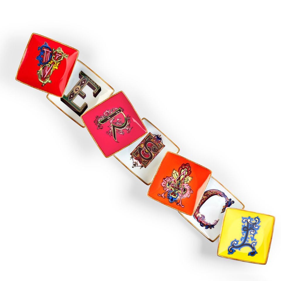 Colorful decorative porcelain trays from Versace’s Alphabet collection, each featuring a different ornate letter in a unique style, spelling out ‘VERSACE’ in a playful arrangement.