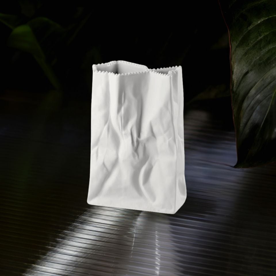 A matte white porcelain vase mimicking a crumpled paper bag, displayed on a dark reflective surface.