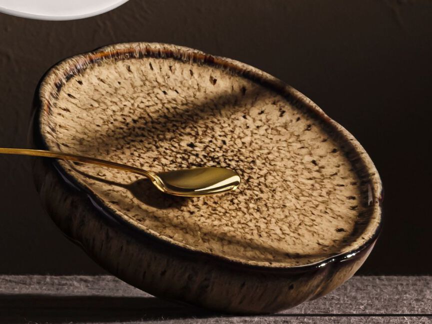 Nahaufnahme eines Rosenthal Tellers mit nat&uuml;rlicher, gesprenkelter Glasur in Beige- und Braunt&ouml;nen. Ein goldfarbener L&ouml;ffel liegt auf dem Teller und betont die hochwertige Materialit&auml;t sowie das moderne, von organischen Formen inspirierte Design