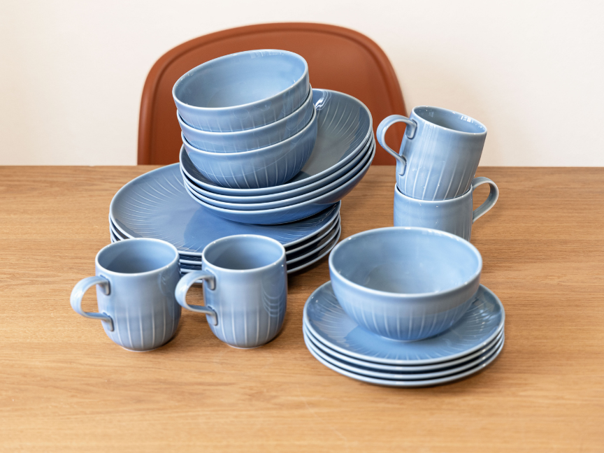 A Rosenthal tableware set in light blue, arranged on a wooden table. Plates, bowls and cups with a delicate embossed pattern are stacked to form a harmonious ensemble, showcasing modern design, high-quality craftsmanship and timeless tableware.